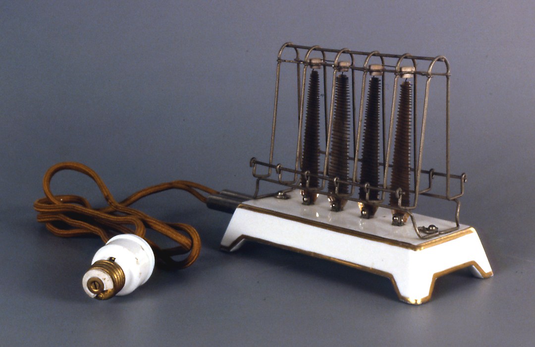 Vroege elektrische broodrooster op een witte porseleinen voet met gouden rand. Daarboven een open frame van metaaldraad rond vier verwarmingselementen. Links een stoffen snoer met een witte lampfitting-stekker.