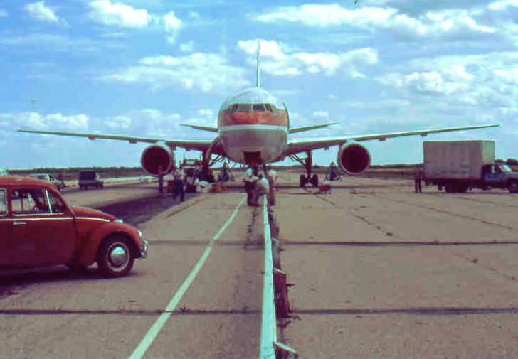 Een Boeing 767 van Air Canada staat van voren gezien op een betonnen landingsbaan, met de neus omlaag op het asfalt. Midden over de baan loopt een vangrail. Op de voorgrond links een rode Volkswagen Kever, rechts een vrachtwagen; rond het toestel staan mensen.